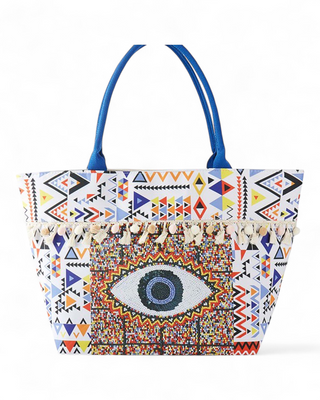 Large white canvas tote with geometric Aztec print, beaded evil eye graphic center, pom-pom tassel trim, and blue handles.
