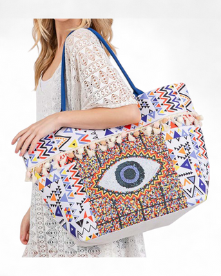 Large white canvas tote with geometric Aztec print, beaded evil eye graphic center, pom-pom tassel trim, and blue handles.