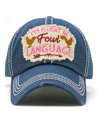 A distressed denim blue baseball cap featuring an embroidered patch with the text "I'm Fluent in Fowl Language" and chicken graphics, offering a playful and humorous accessory.