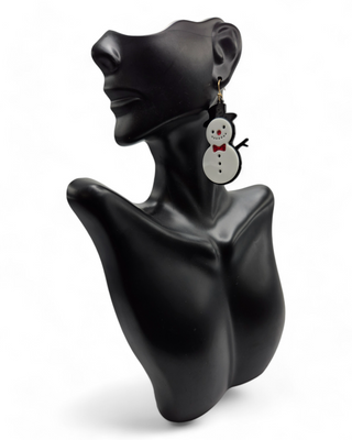 Snowman-shaped dangle earrings worn on a black mannequin bust. The snowmen feature glittery red bow ties and noses, with black top hats and arms, creating a whimsical holiday look.